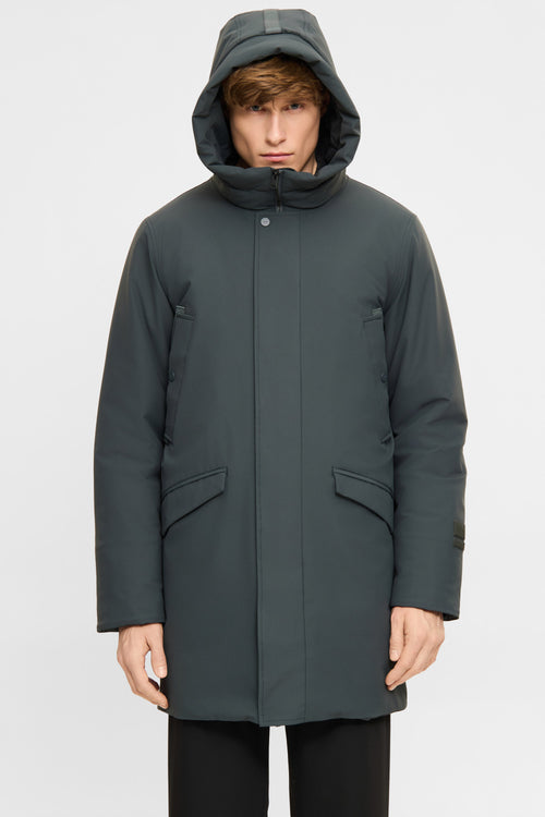 A man wearing a dark gray hooded parka jacket with a high collar and front flap pockets, photographed against a light gray studio background.
