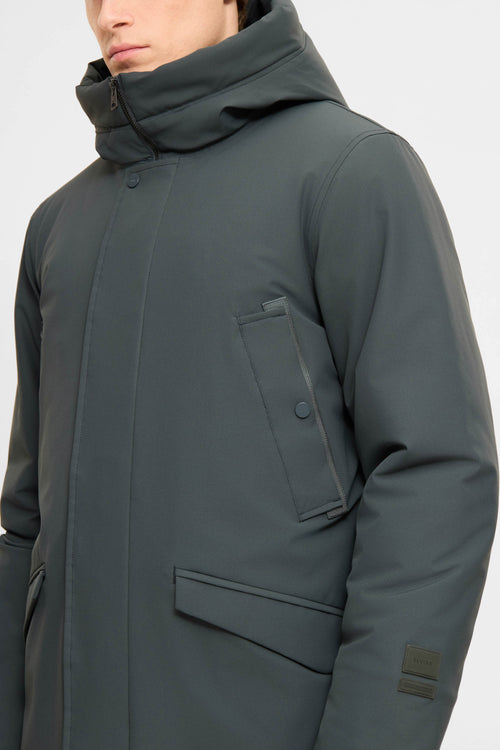 A close-up of a person wearing a dark gray hooded jacket with a high collar, front flap pocket, and a small rectangular patch on the sleeve, photographed against a light gray background.