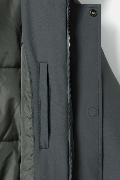 Close-up of a dark gray jacket showing the front zipper, snap button, vertical welt pocket, and quilted puffer lining.