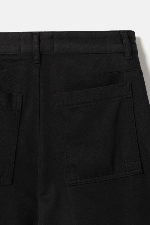 Close-up of the back waistband and right rear pocket of black trousers, showing belt loops, stitching, and a small tonal embroidered logo near the pocket corner.