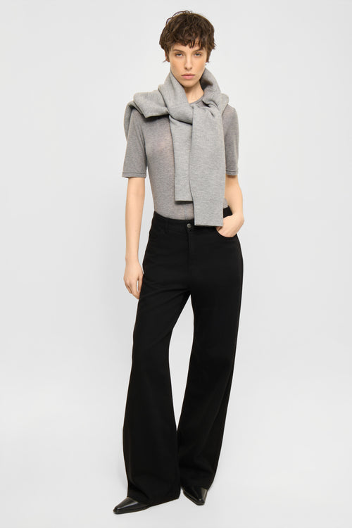 A model stands against a light gray studio background wearing a light gray knit wrap scarf draped over a short-sleeve top and black wide-leg trousers.