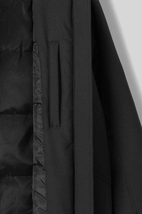 Close-up detail shot of a black puffer jacket, showing quilted padding, a vertical seam, and a zippered pocket on a plain light gray background.